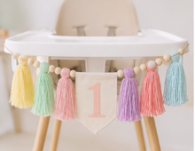 Load image into Gallery viewer, A pastel tassel banner garland with wooden beads and a central fabric pennant marked with the number 1, hanging across a white high chair.