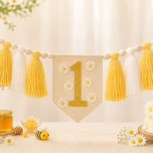 Load image into Gallery viewer, A honey-themed first birthday party decoration set featuring a burlap banner with a gold glitter number 1 and white daisy flowers, hanging alongside yellow and white yarn tassels. The scene is styled with a white daisy cake, honey jars, a small bee toy, and daisy flowers on a light wooden table.
